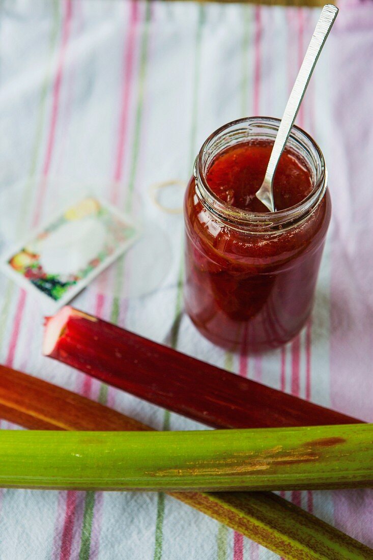 A jar of strawberry and rhubarb jam with fresh rhubarb, labels and utensils