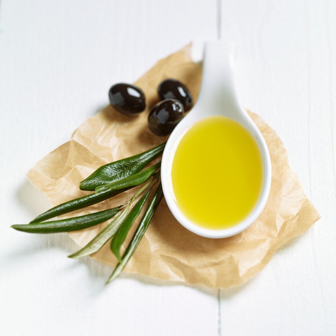 Ein Löffel Olivenöl, Olivenblätter und schwarze Oliven auf Papier