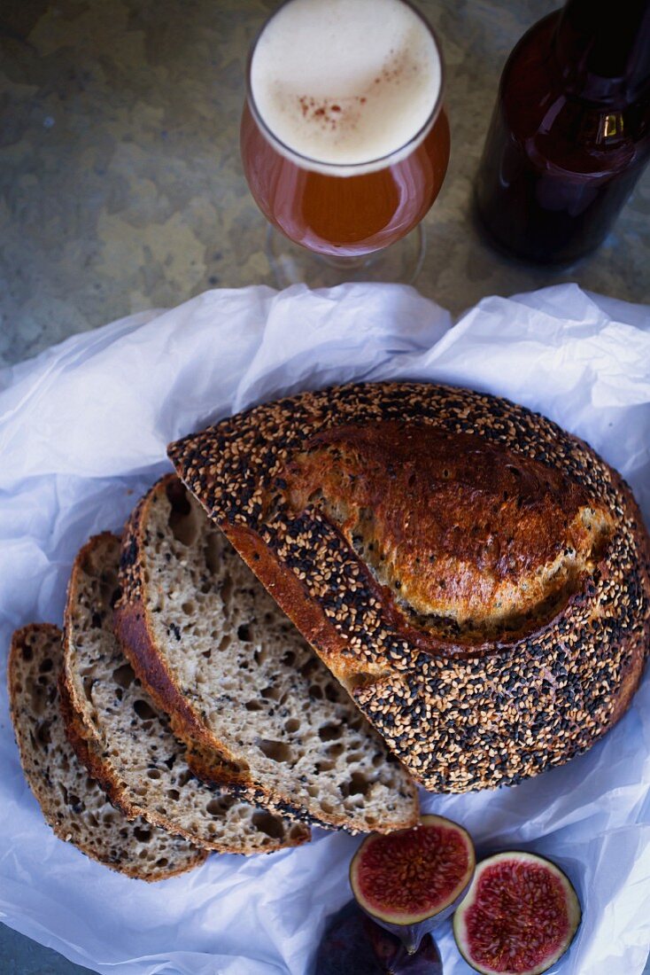 A sourdough loaf with a sesame seed crust, sliced, next to a beer and fresh figs