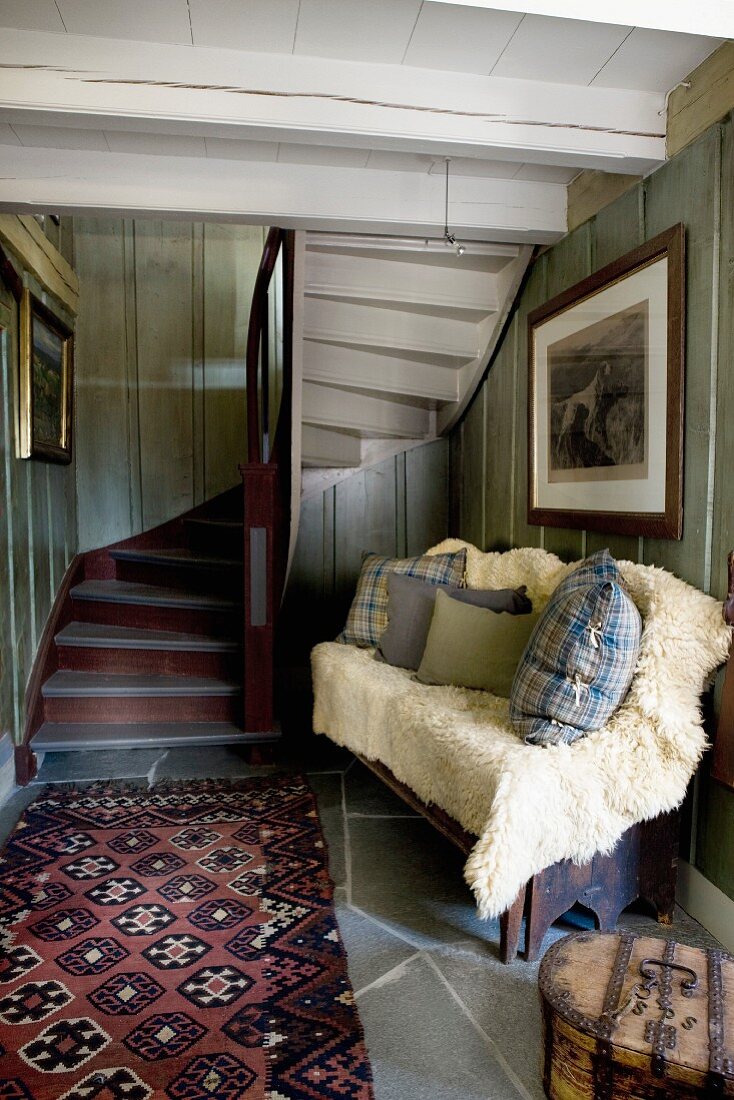 Cushions and pale fur blanket on bench next to rug at foot of winding wooden staircase in rustic foyer