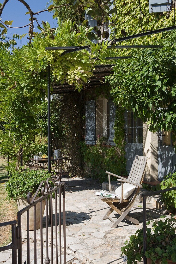 Kleine Steinterrasse mit berankter Eisenpergola vor provenzalischem Gästehaus mit klassisch hellblauen Fensterläden