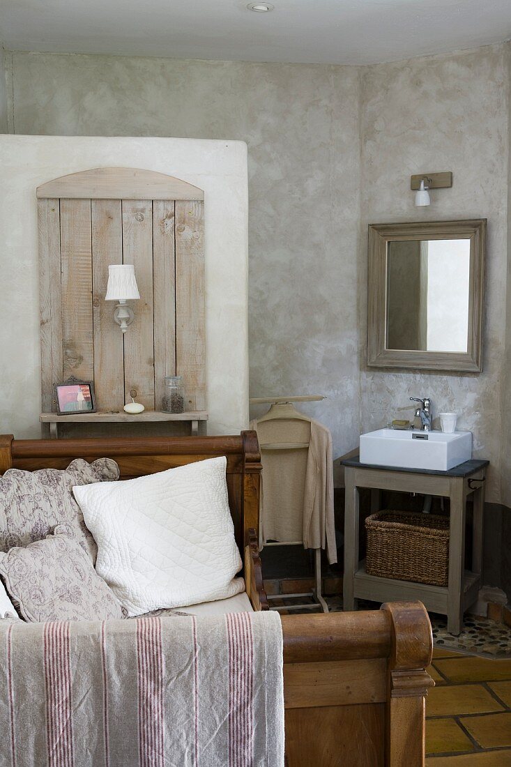Antikes Bett mit hölzernen Seitenteilen, Wandelement mit Leuchte und Ablage auf Trennmauer zur Toilette, daneben ein schlichter Waschtisch