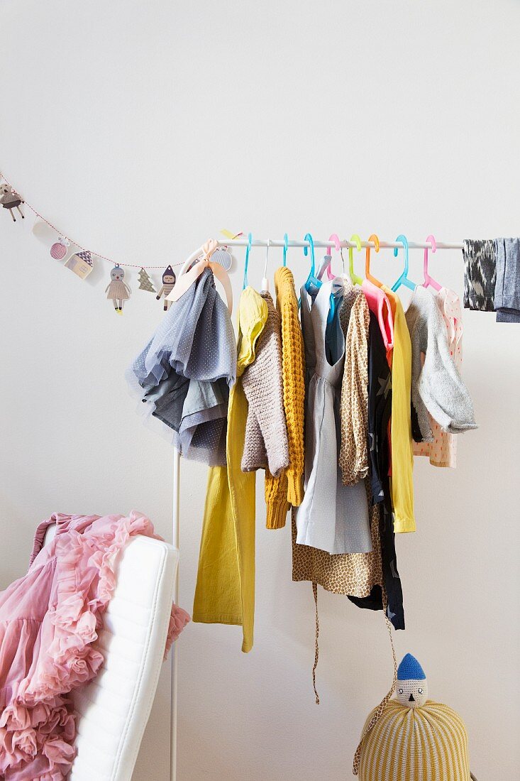 Children's clothing on colourful, plastic coat hangers on rail and pink, ruched skirt draped over chair back