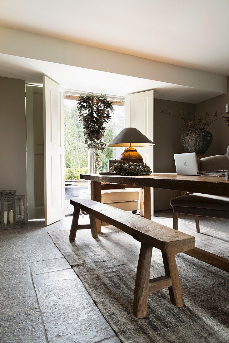 Rustic wooden table and bench set in modern interior with open interior shutters on terrace door in background