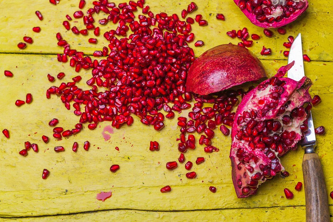 Pomegranates, whole and chopped, surrounded by seeds on a rustic yellow wooden surface