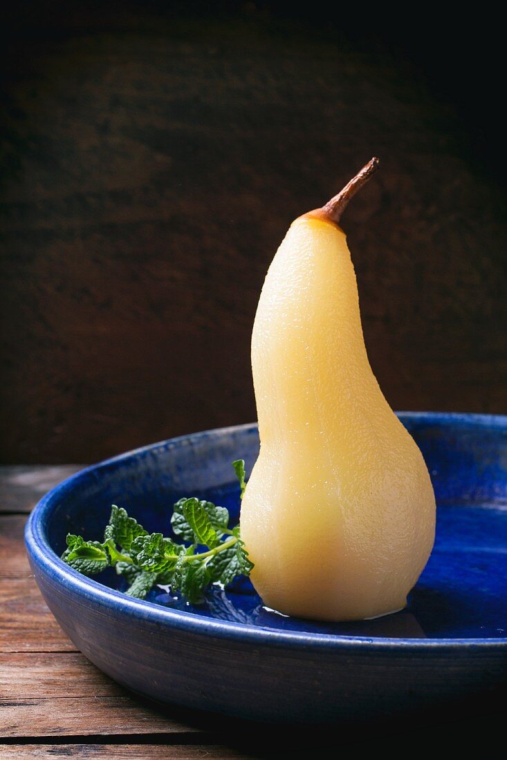 A pear poached in white wine with syrup and fresh mint in a blue ceramic bowl