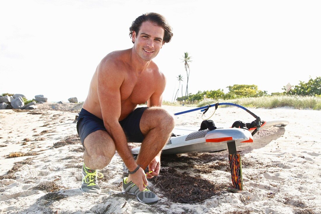 A young, topless man wearing shorts tying his shoes on a beach