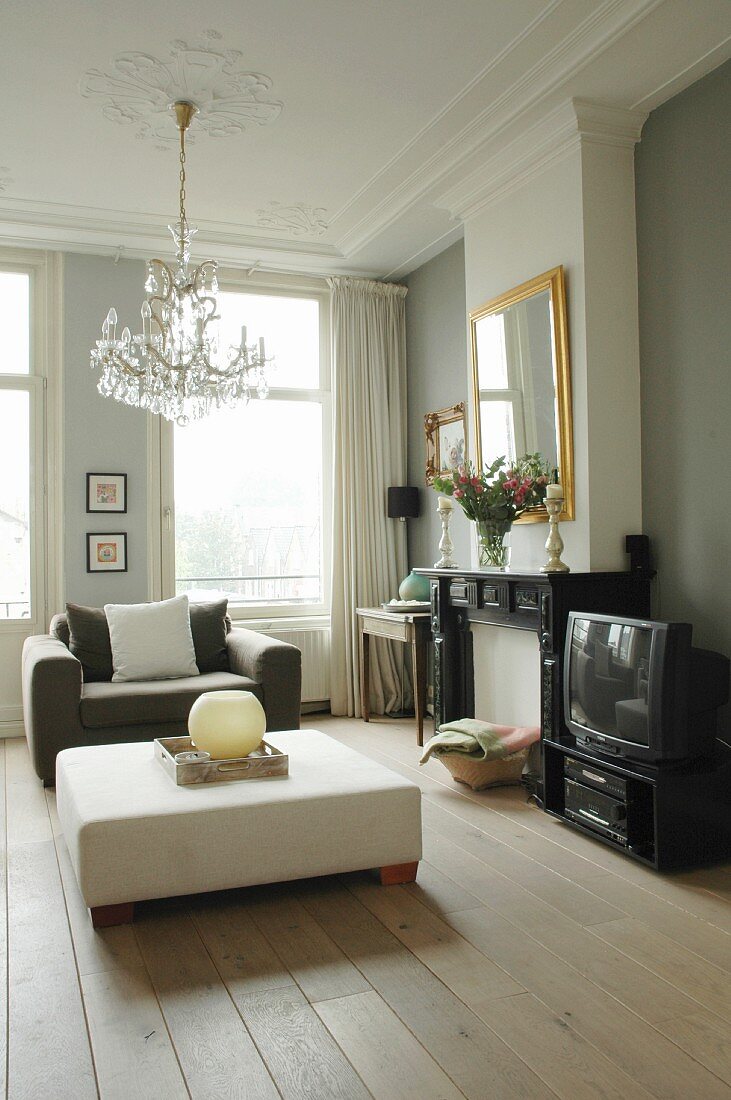 Grand living room with minimalist ambiance with low, white ottoman and grey armchair below chandelier hanging from stucco ceiling