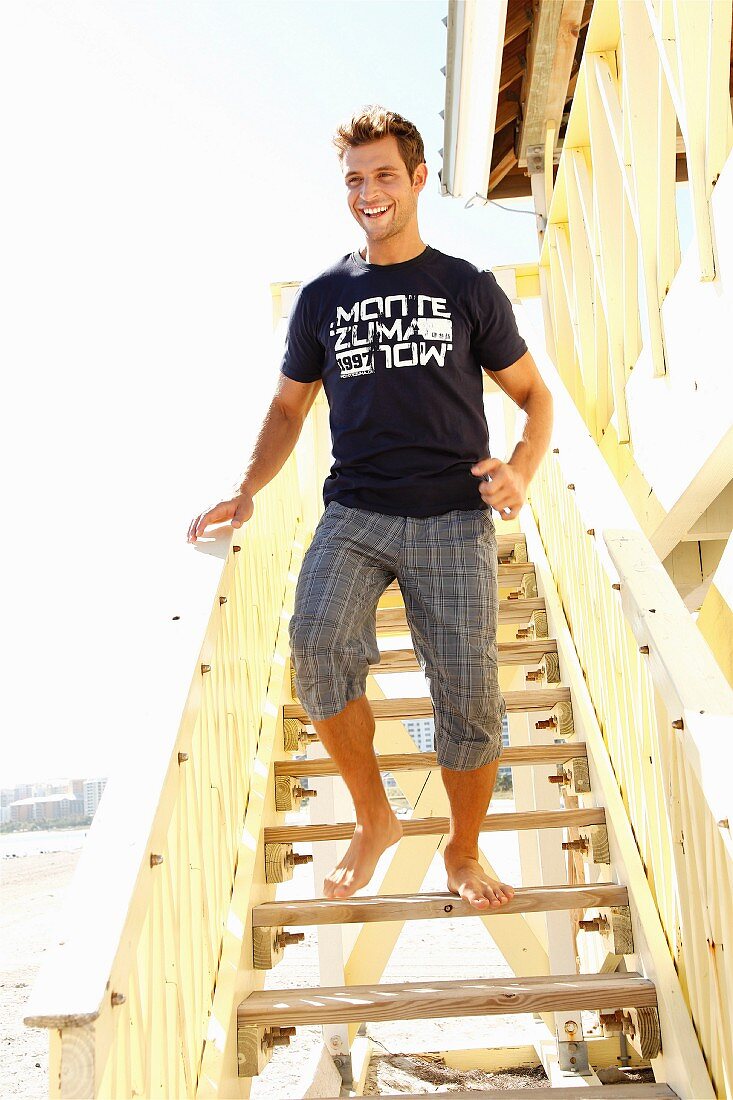 A young man on a flight of wooden step on a beach wearing a t-shirt and a checked trousers