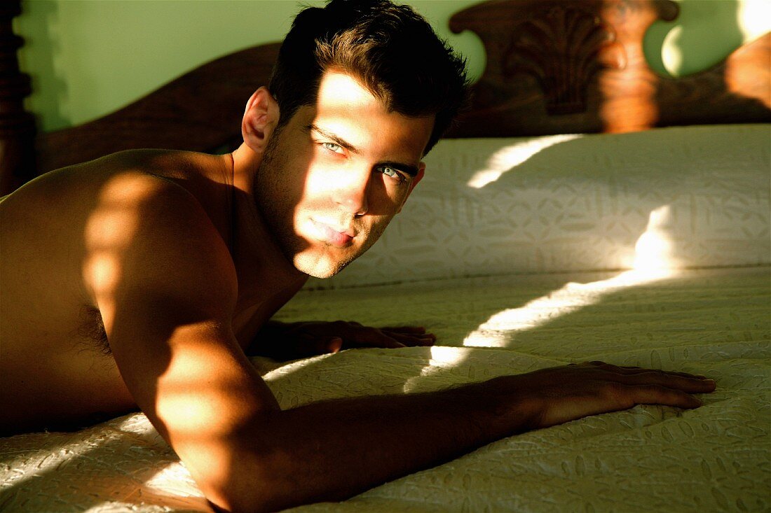 A young, topless man lying on a bed in light and shade