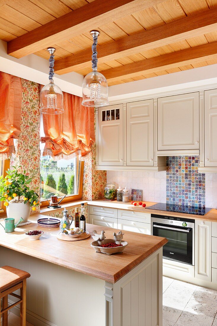 Pale, country-house-style kitchen with breakfast bar below pendant lamps hanging from wooden ceiling