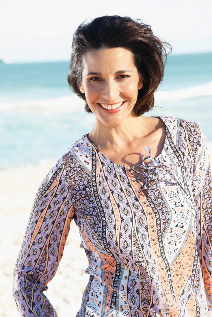 A dark-haired woman on a beach wearing a patterned blouse