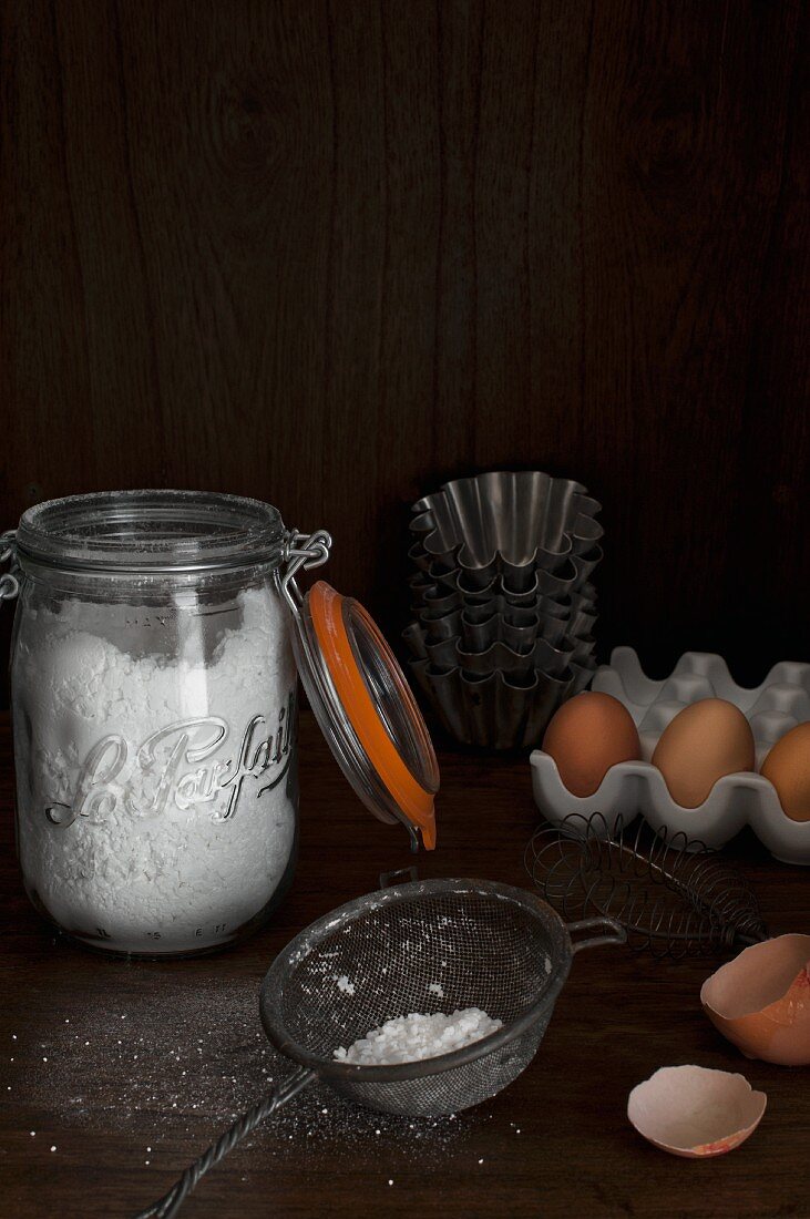 A jar of flour, a metal sieve, eggs and baking tins