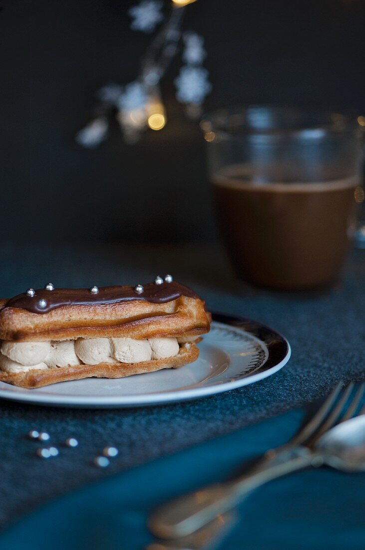 Eclair mit Schokoladenglasur, Silberperlen und Kaffeecreme vor einer Kaffeetasse (weihnachtlich)