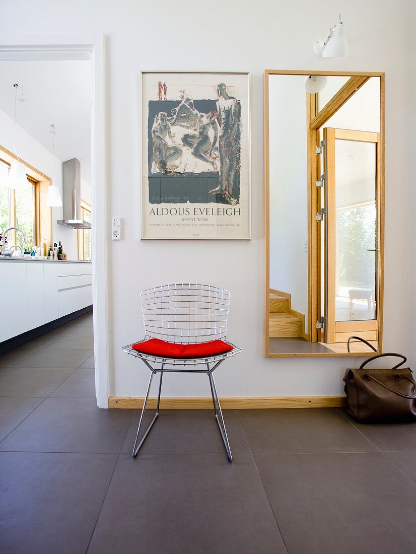 A classic, mesh metal chair and a red cushion in front of a full-length mirror and door at the side open to the kitchen with continuous dark grey tiled floor