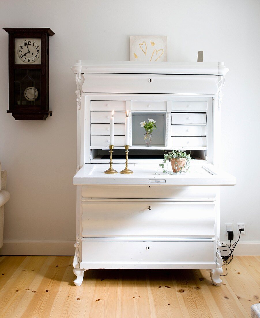 White-painted bureau with lit candles in brass candlesticks on folded-down desk in rustic interior