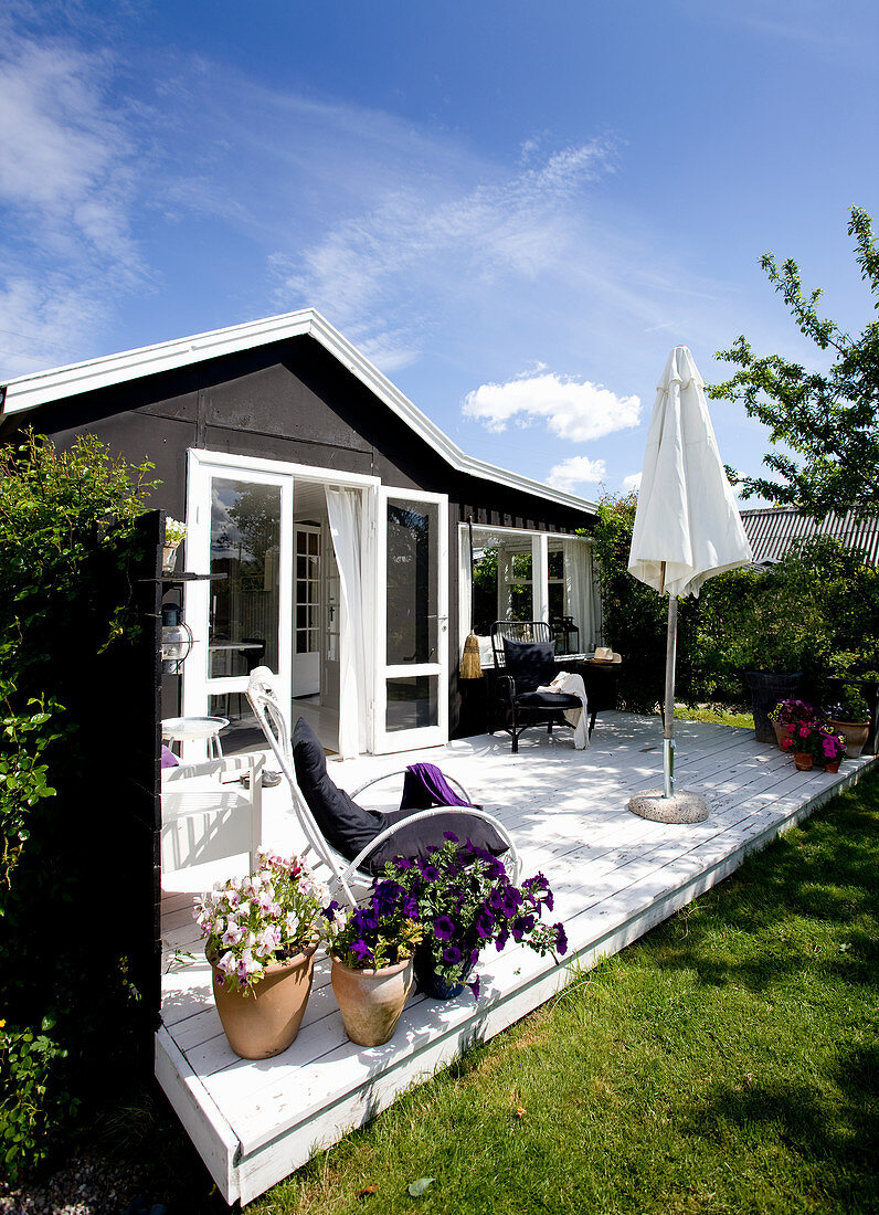 Weiß lackierter Bambusstuhl neben Stiefmütterchen und Petunien auf sonniger Terrasse vor Sommerhaus mit weissen Fensterrahmen und dunkler Fassade