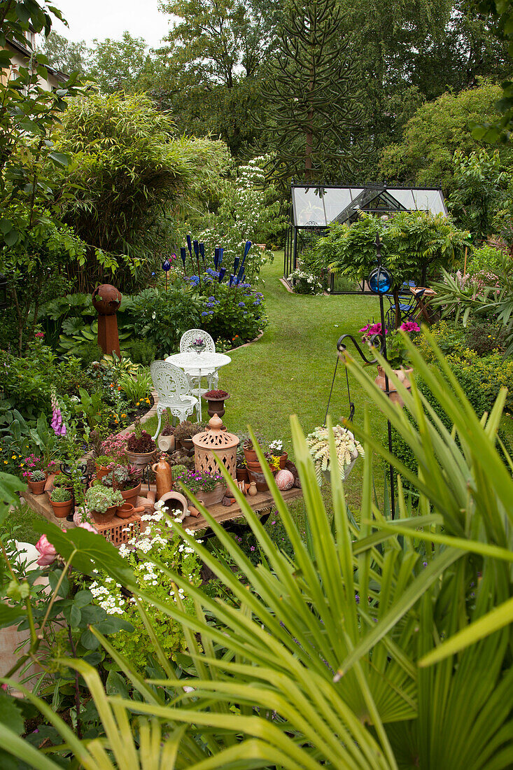 Lawn flanked by densely planted flowerbeds with bright blue glass ornaments and potted succulents on table; palm fronds in foreground