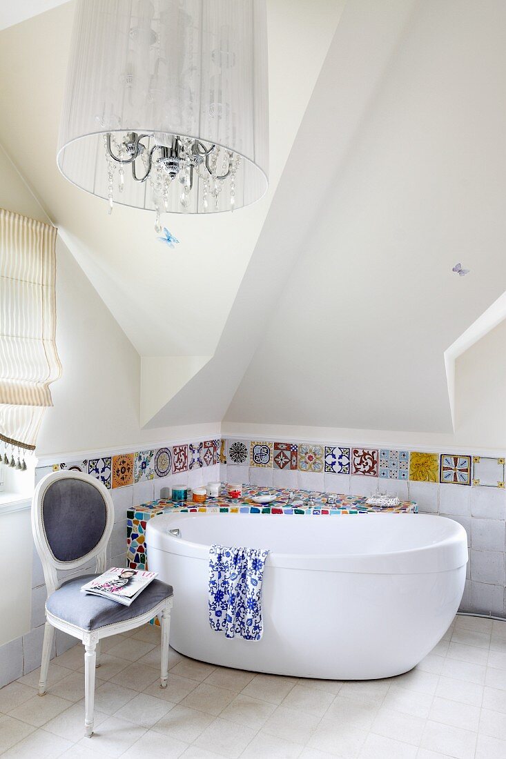 Surface covered in mosaic of colourful tile shards and tiled border behind free-standing bathtub and antique chair; chandelier with lampshade in foreground