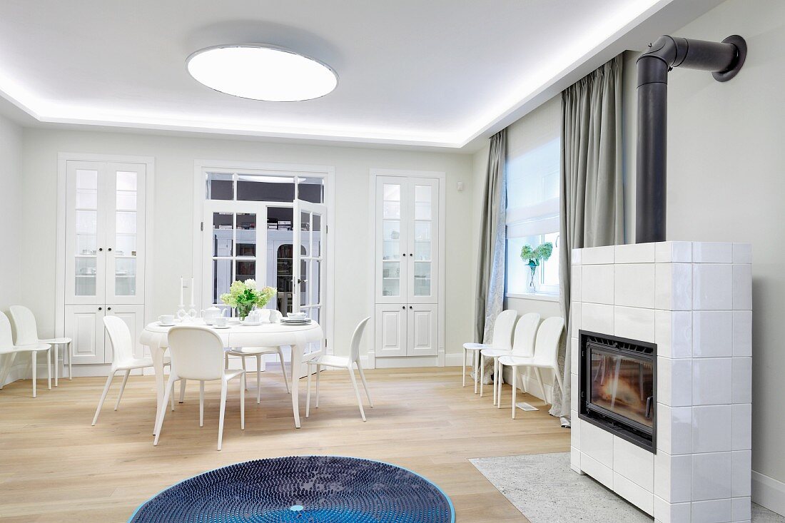 Dining set and extra chairs against wall in white interior; blue rug in front of tiled wood-burning stove in foreground