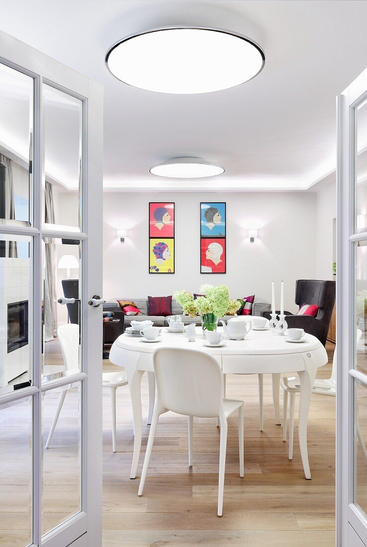 View through open glass double doors of white dining area with plastic chairs and pop-art pictures above seating area in background