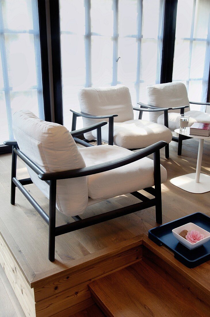 Elegant armchair with black frame and white cushions and bowl of flowers on tray on wooden platform next to glass wall