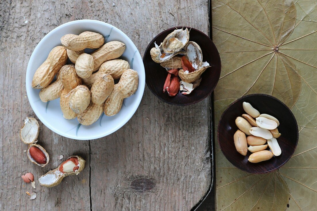 Erdnüsse, geschält und ungeschält, in verschiedenen Schälchen auf rustikalem Brett
