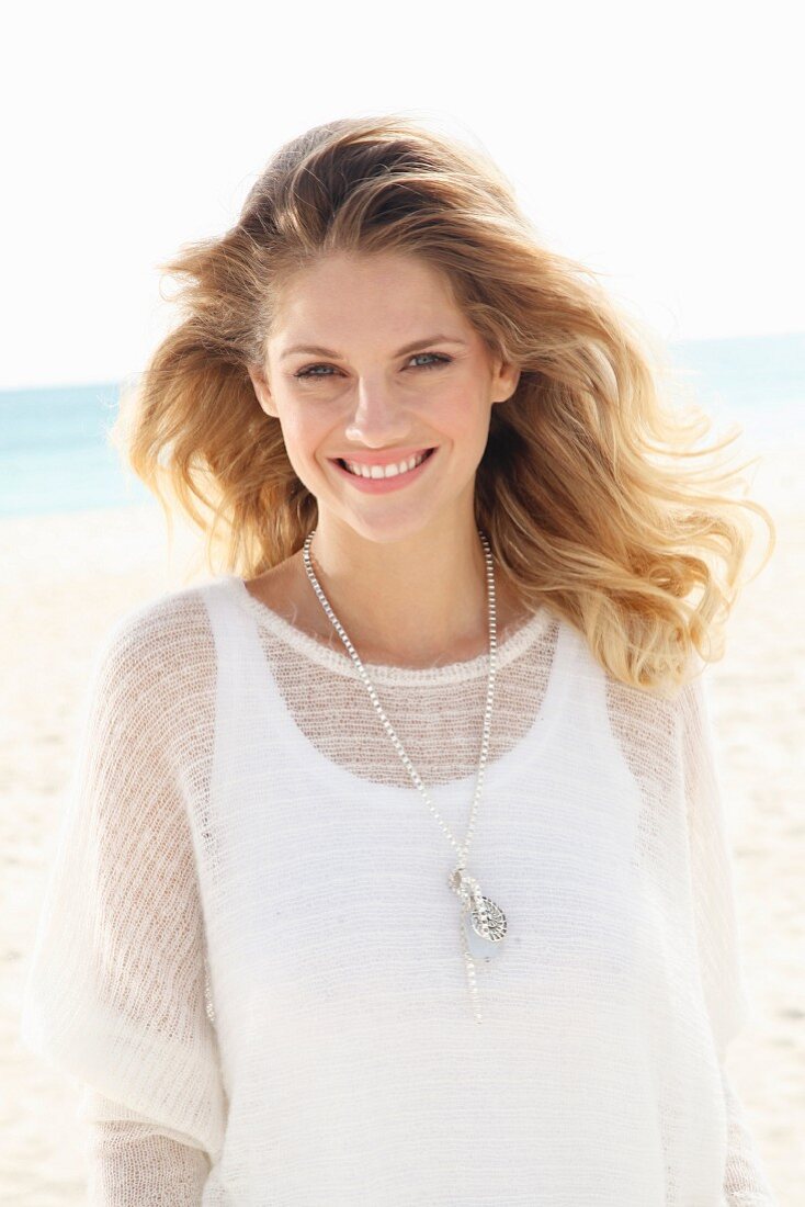 Junge blonde Frau in weißem Top und transparentem Strickpulli steht am Strand