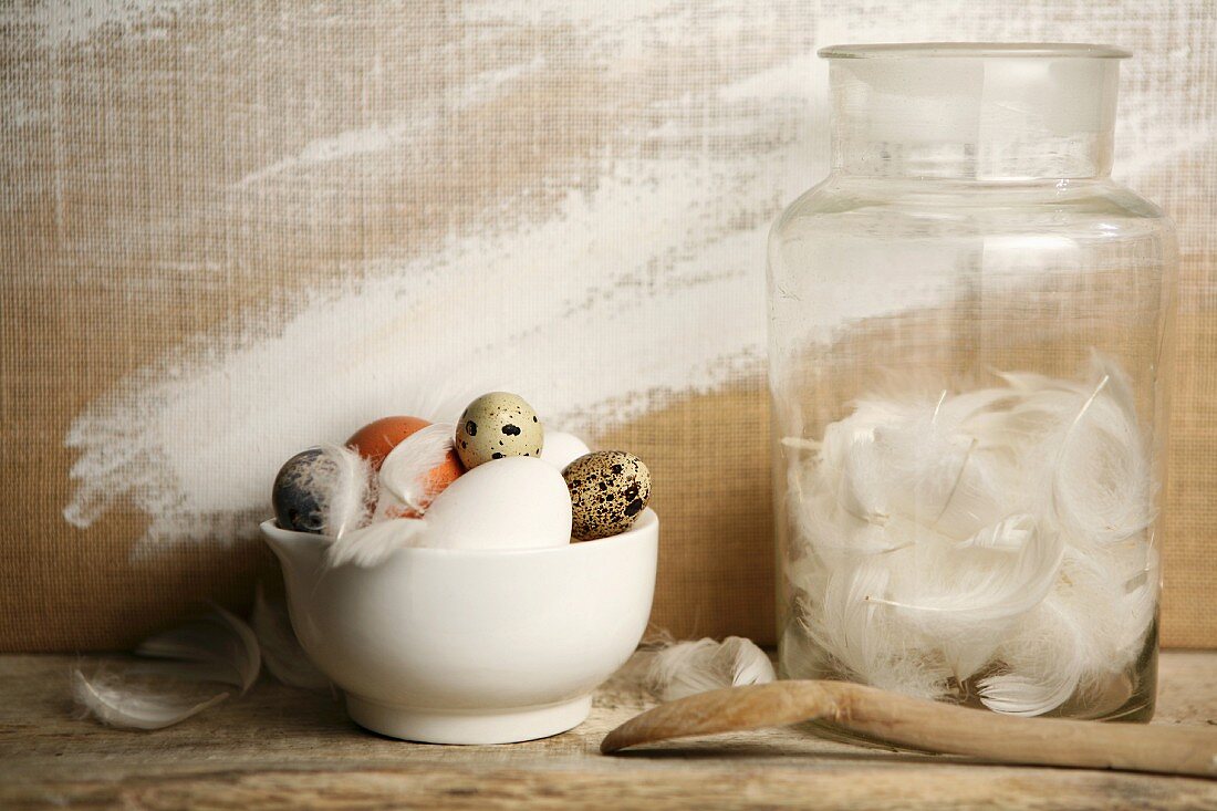 Various eggs in a bowl next to a jar of white feathers