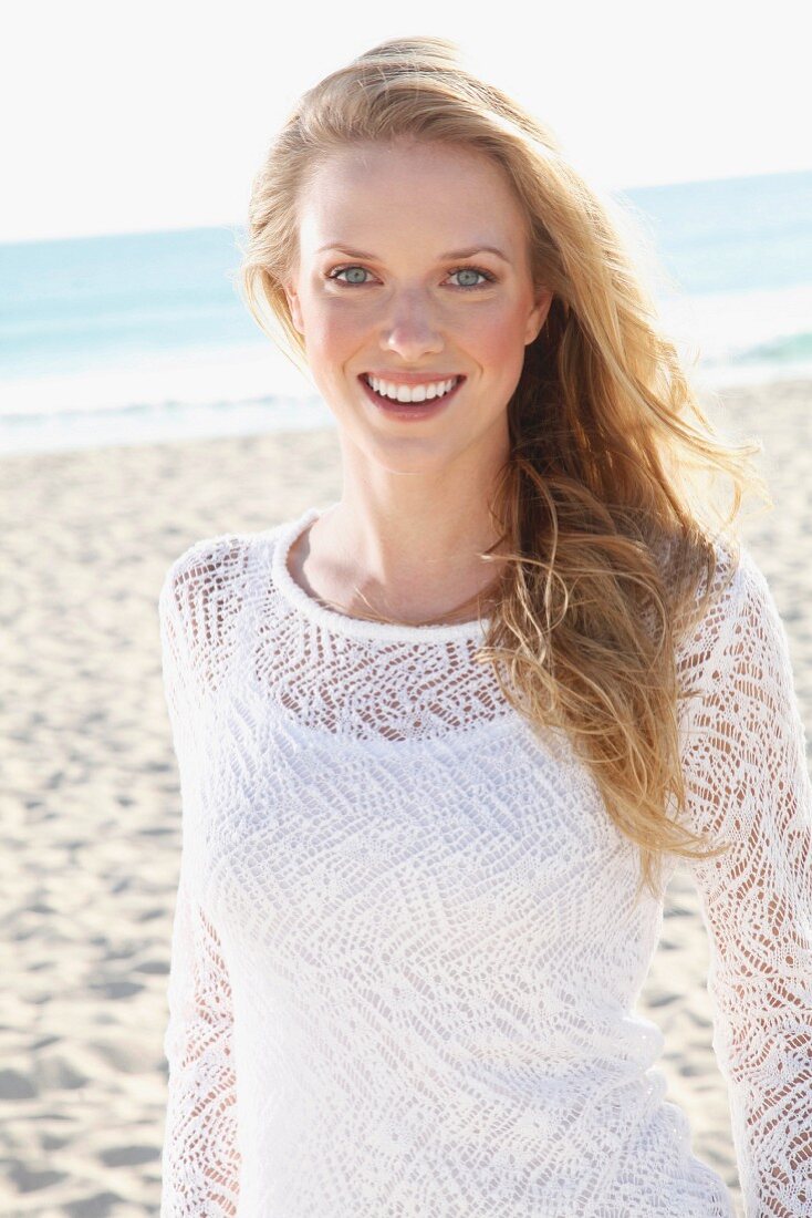 Junge blonde Frau in weißem Top und transparentem Strickpulli am Strand