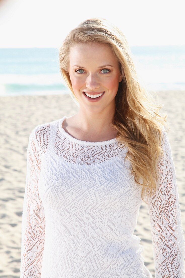 A young blonde woman on a beach wearing a white top and a transparent knitted jumper