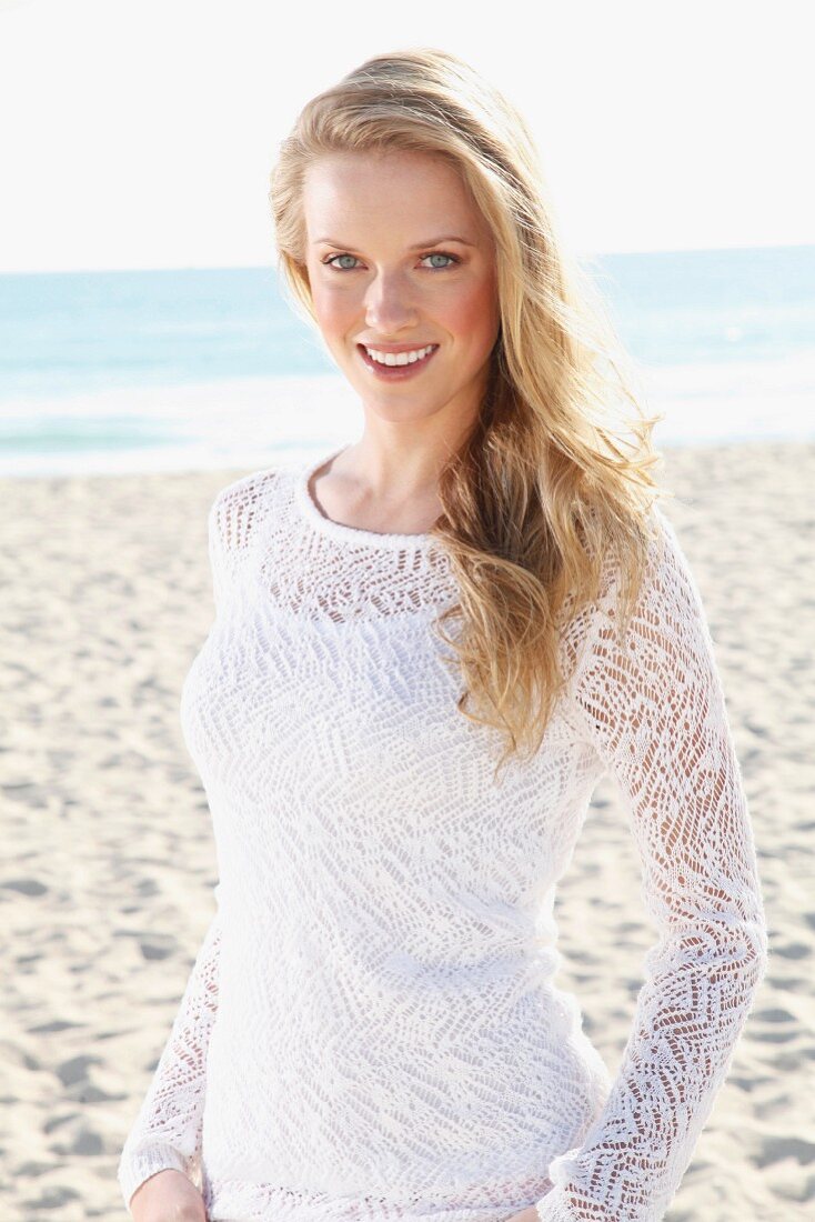 A young blonde woman on a beach wearing a white top and a transparent knitted jumper