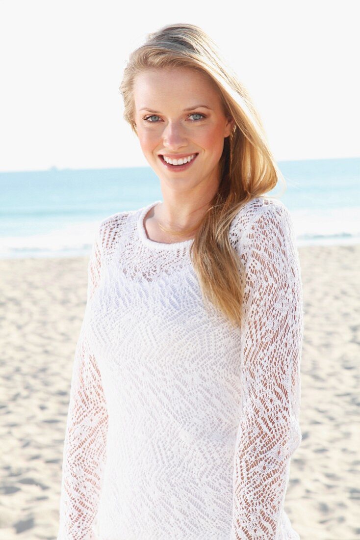 A young blonde woman on a beach wearing a white top and a transparent knitted jumper