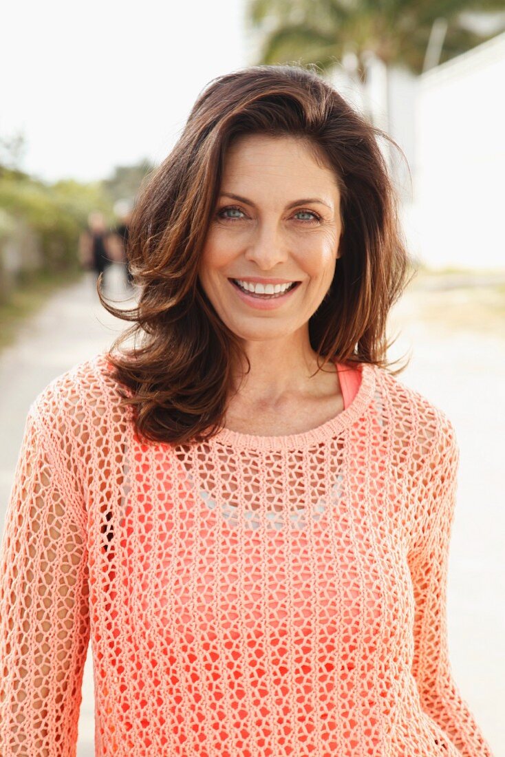 A brunette woman on a beach wearing a salmon-coloured top and an openwork jumper