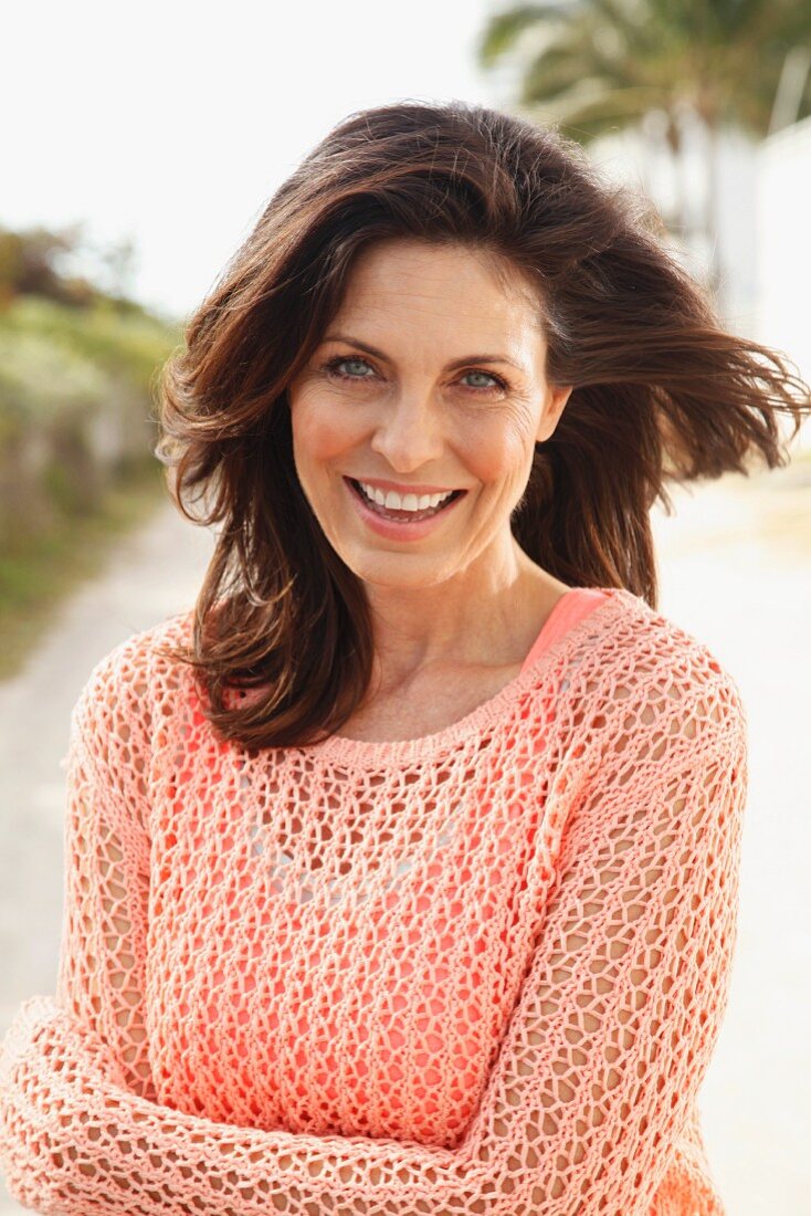 A brunette woman on a beach wearing a salmon-coloured top and an openwork jumper