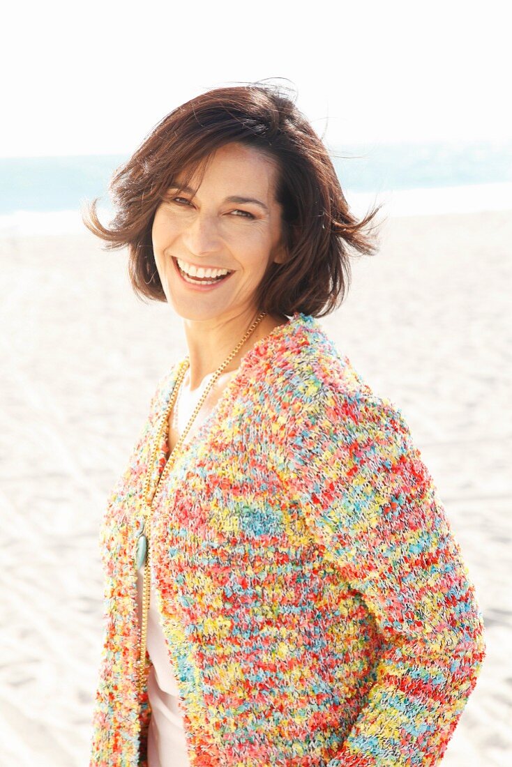 A brunette woman on a beach wearing a white top and colourful cardigan