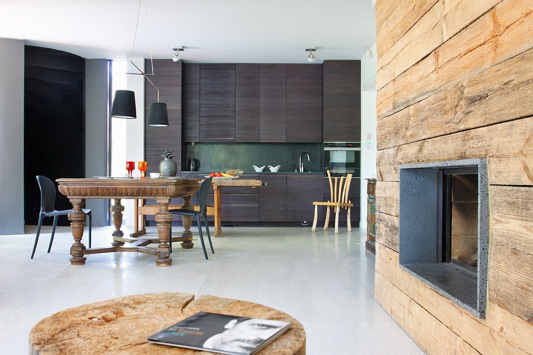 Fireplace in rustic wooden wall; dining area with antique table and modern, charcoal grey kitchen counter in background