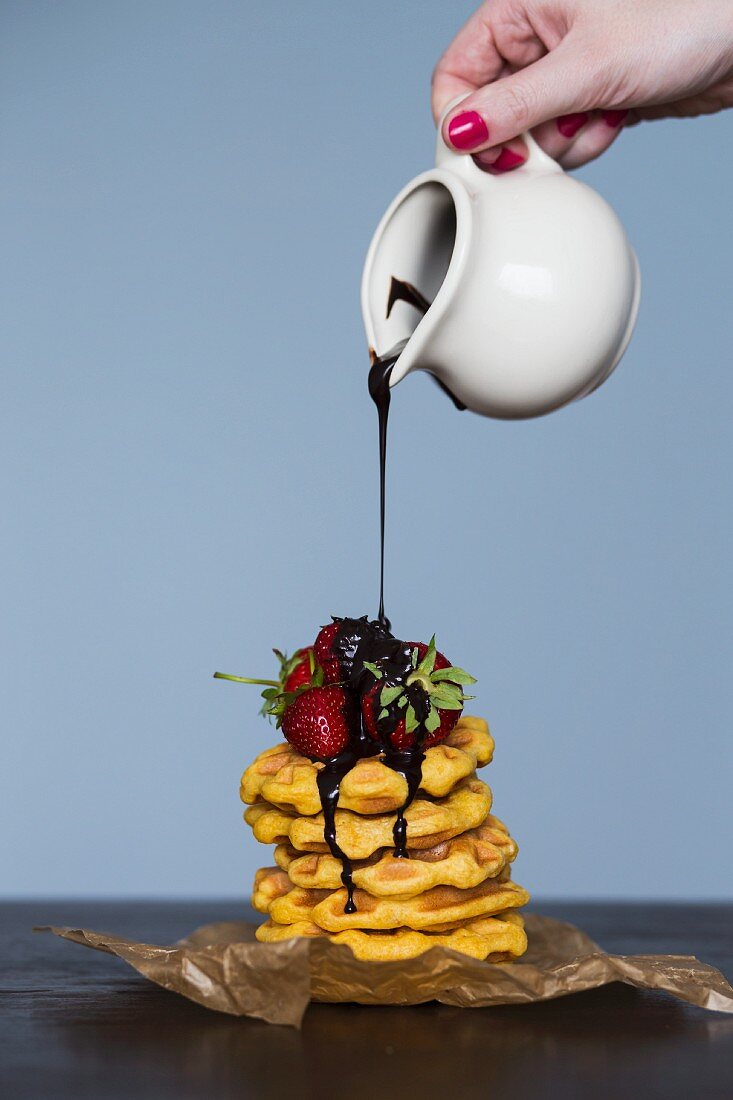 Chocolate sauce being poured over a stack of waffles topped with fresh strawberries