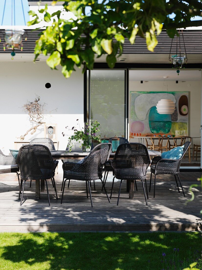 Geflechtstühle in Graubraun um Tisch auf Holzterrasse, vor zeitgenössischem Wohnhaus