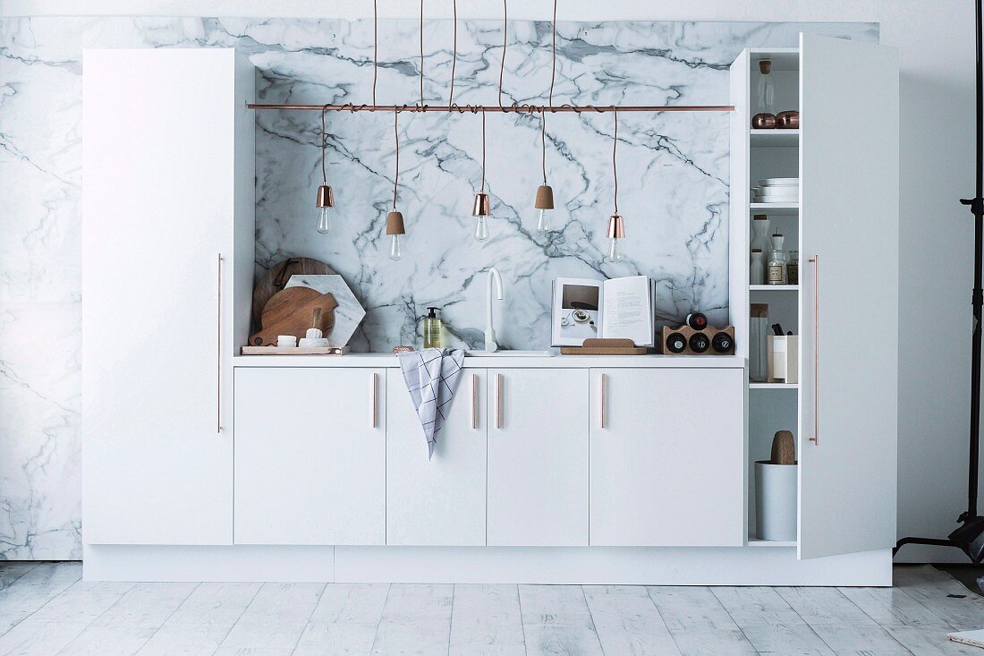 White kitchenette with tall cupboards, integrated counter, row of pendant lamps with cords wrapped around rod and marbled effect on wall
