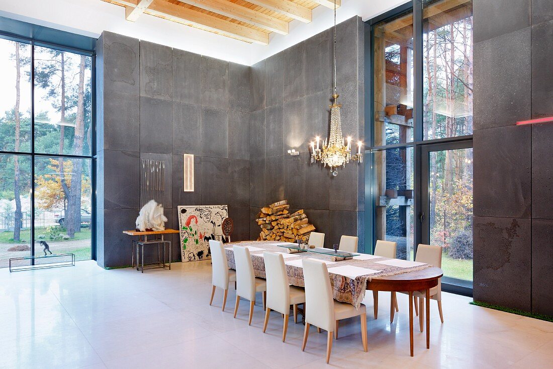 Chandelier above long dining table with upholstered chairs in double-height interior with charcoal-grey wall panels and tall windows