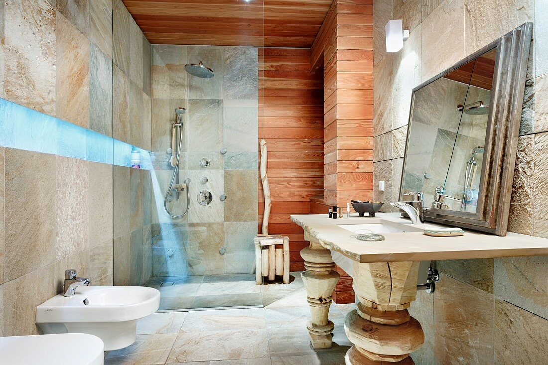 Bathroom with blue lighting strip between stone-tiled walls and horizontal wooden cladding, washstand with carved wooden legs and framed mirror