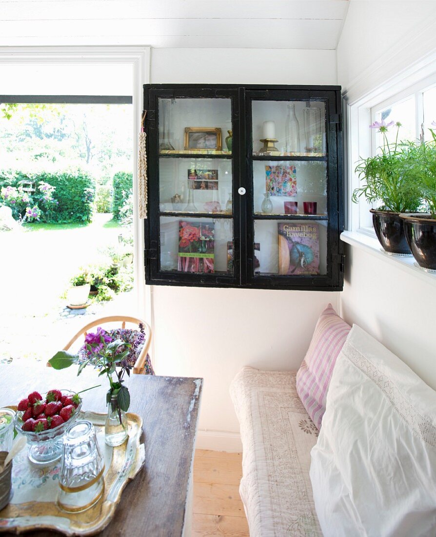 Dining area with bench and small glass-fronted cabinet with dark wooden frame on shelf next to open French windows