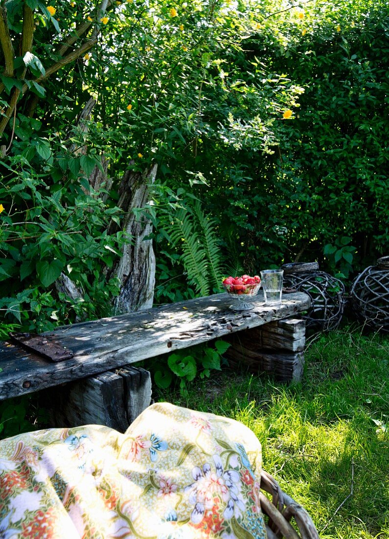 Schattiges Gartenplätzchen mit verwitterter Holzbank, davor gemusterte Picknickdecke im Garten