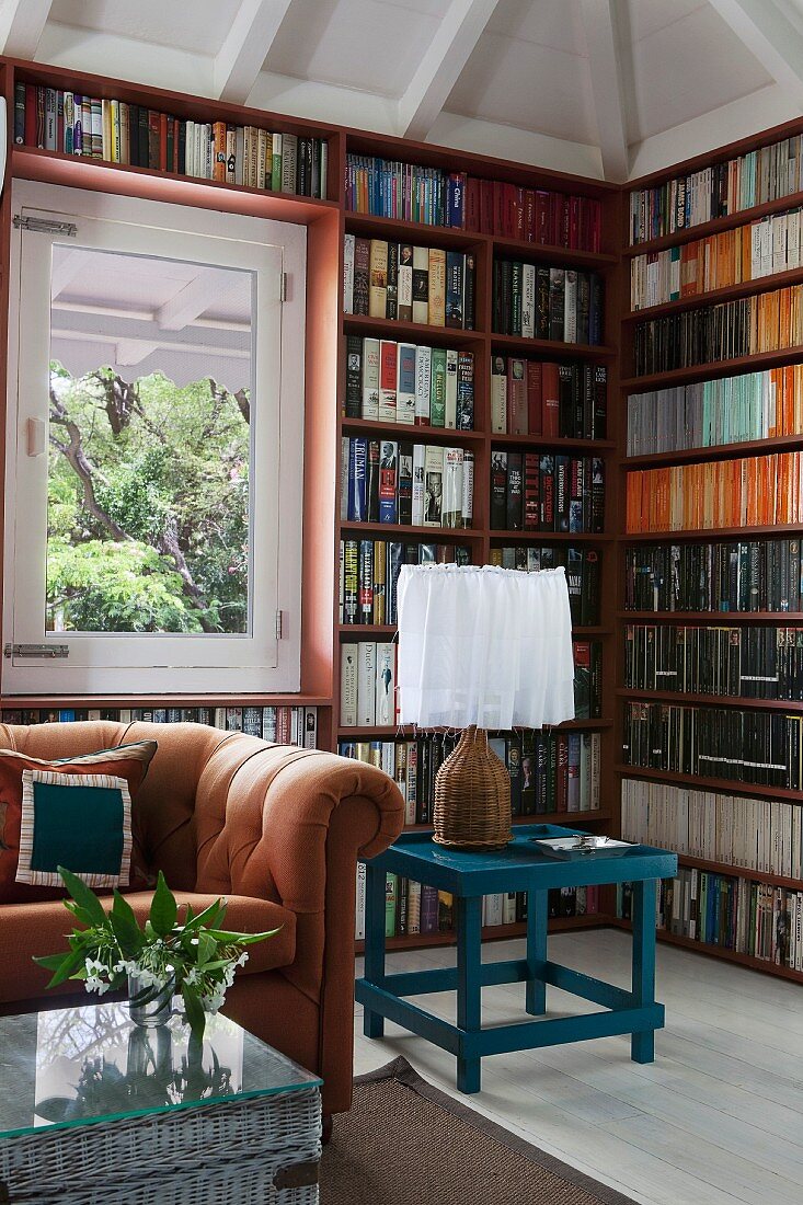 Sitzplatz mit karamellfarbenem Sofa, Korbkiste und petrolfarbenem Tisch vor umlaufend eingebauten Bücherregalen