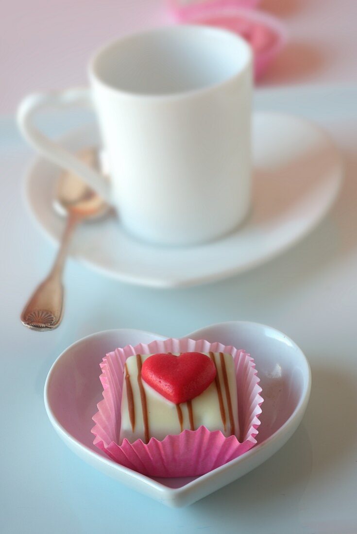 A petit four decorated with a heart in front of a mocha cup