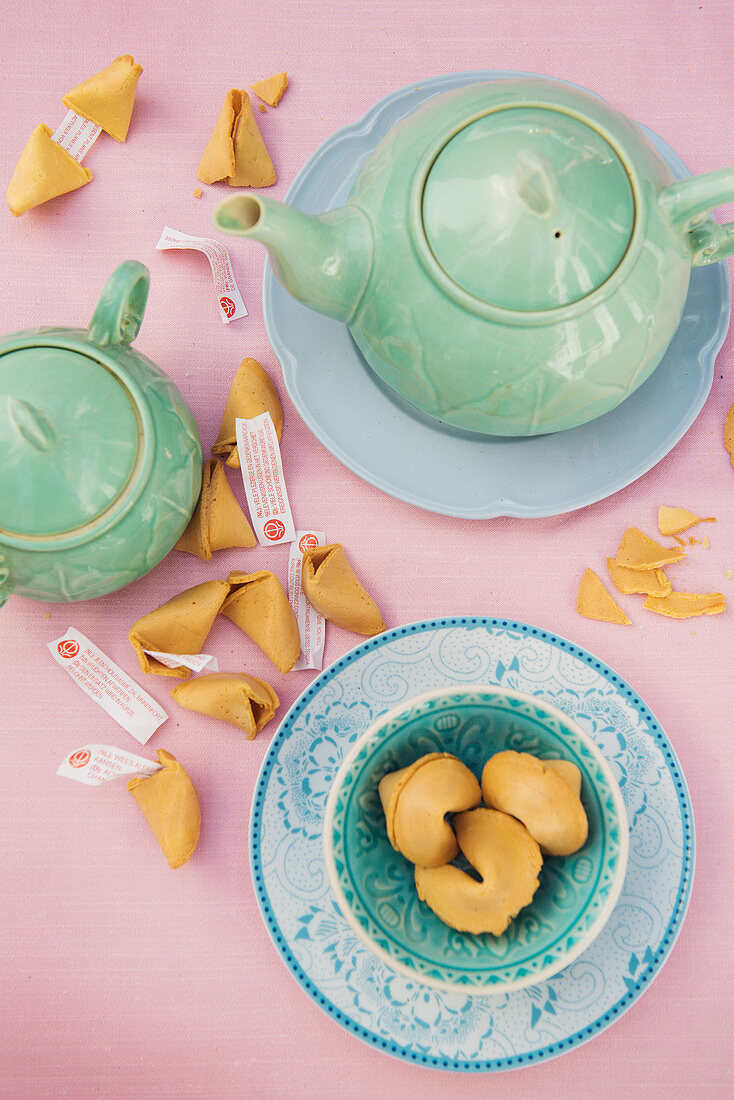 Pastellblaues Teegedeck, Teekanne und aufgebrochene Glückskekse auf rosa Tischdecke