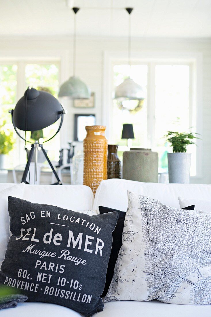 Scatter cushions with various motifs on white sofa in front of collection of vessels and table lamp