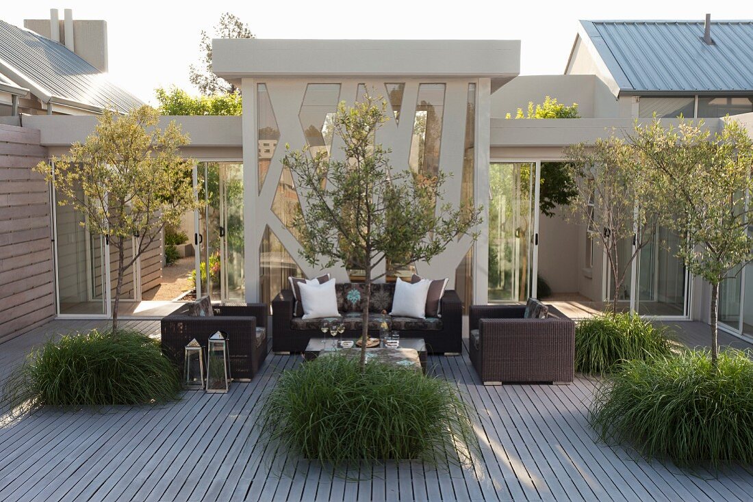 Terrasse mit Bäumen in Trögen, im Hintergrund moderne Outdoor Möbeln aus dunklem Rattan vor Stahl-Glasfassade