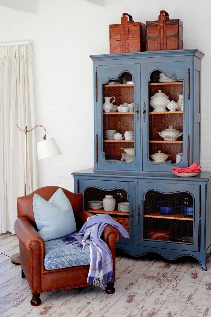 Old-fashioned crockery in blue vintage dresser and brown leather armchair on old wooden floor